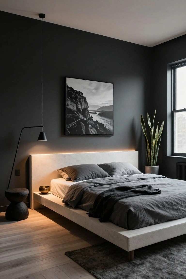 Modern bedroom interior with dark gray walls, low white platform bed featuring LED strip lighting on headboard, gray linens and throw, large black-and-white framed landscape photograph, potted snake plant near window, black floor lamp, pendant light, and light oak wood floors with rug.