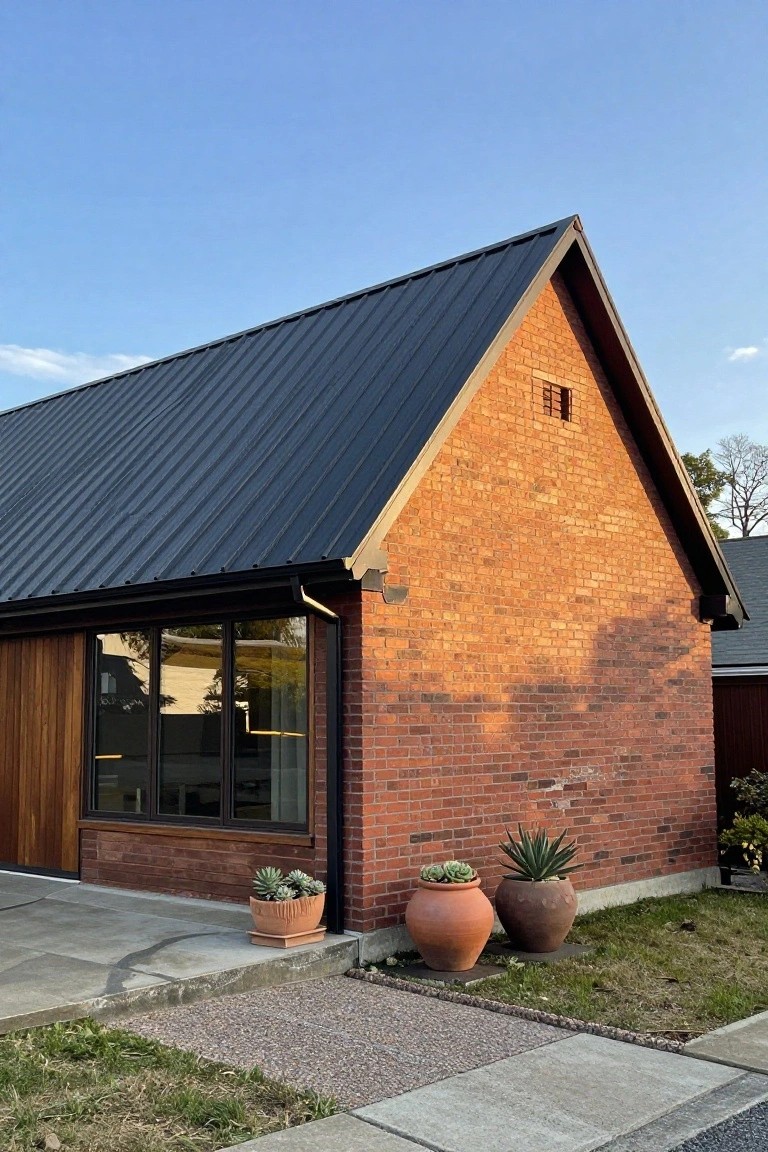 Brick house exterior with black standing seam metal gabled roof, large horizontal wood-framed windows, terracotta pots with succulents and agave plants on concrete slab near gravel path.