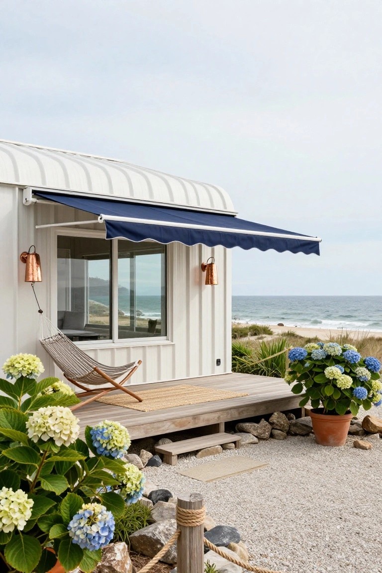 White corrugated metal house with extended navy blue scalloped awning over a window and wooden deck porch, hammock chair, potted hydrangeas, rocks, gravel ground, and ocean view in the background.