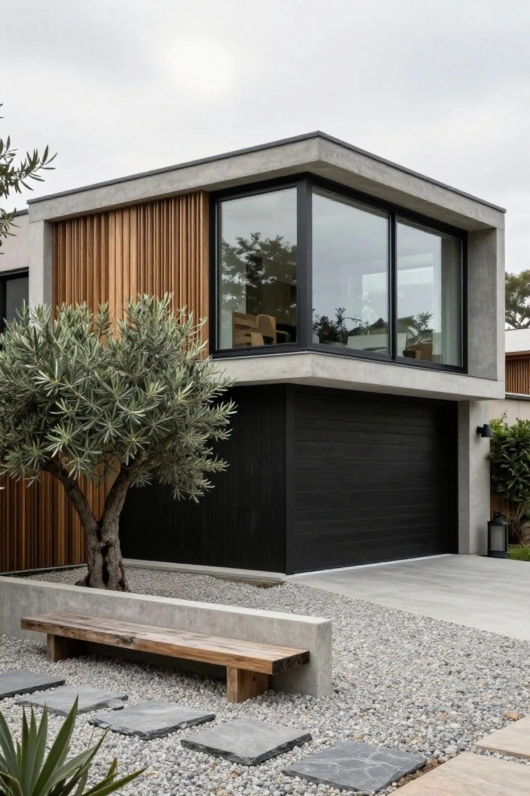 Corner view of a modern two-story house with gray concrete walls, vertical light wood cladding on one upper section beside large black-framed windows, black garage door, olive tree in front, concrete bench, gravel ground cover, and irregular stone stepping stones.