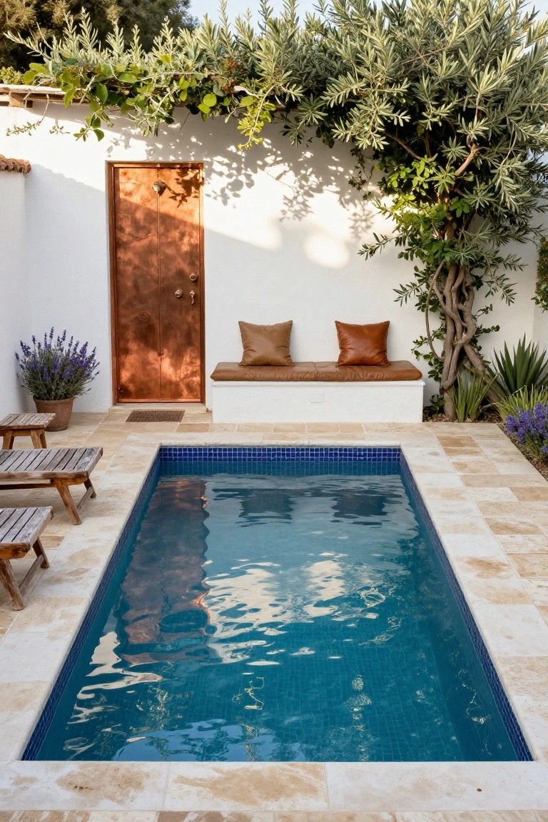 Narrow rectangular pool with blue mosaic tiles in a travertine-paved courtyard, next to a white wall with copper door, built-in bench with tan leather cushions, lavender pots, wooden lounge chairs, and greenery climbing the walls.