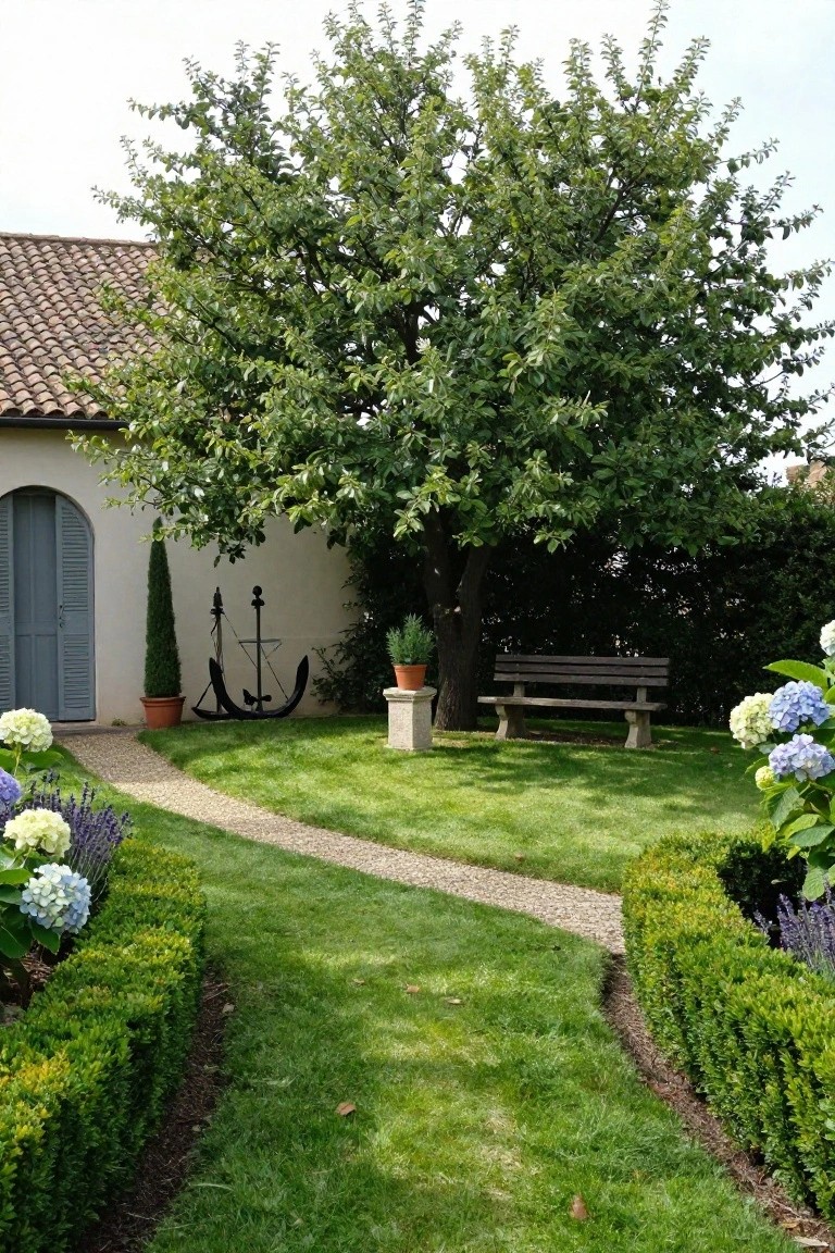Winding gravel path bordered by low clipped boxwood hedges through a green lawn, flanked by hydrangeas and lavender, with a wooden bench under a tree near a white stucco house wall featuring a metal anchor sculpture.
