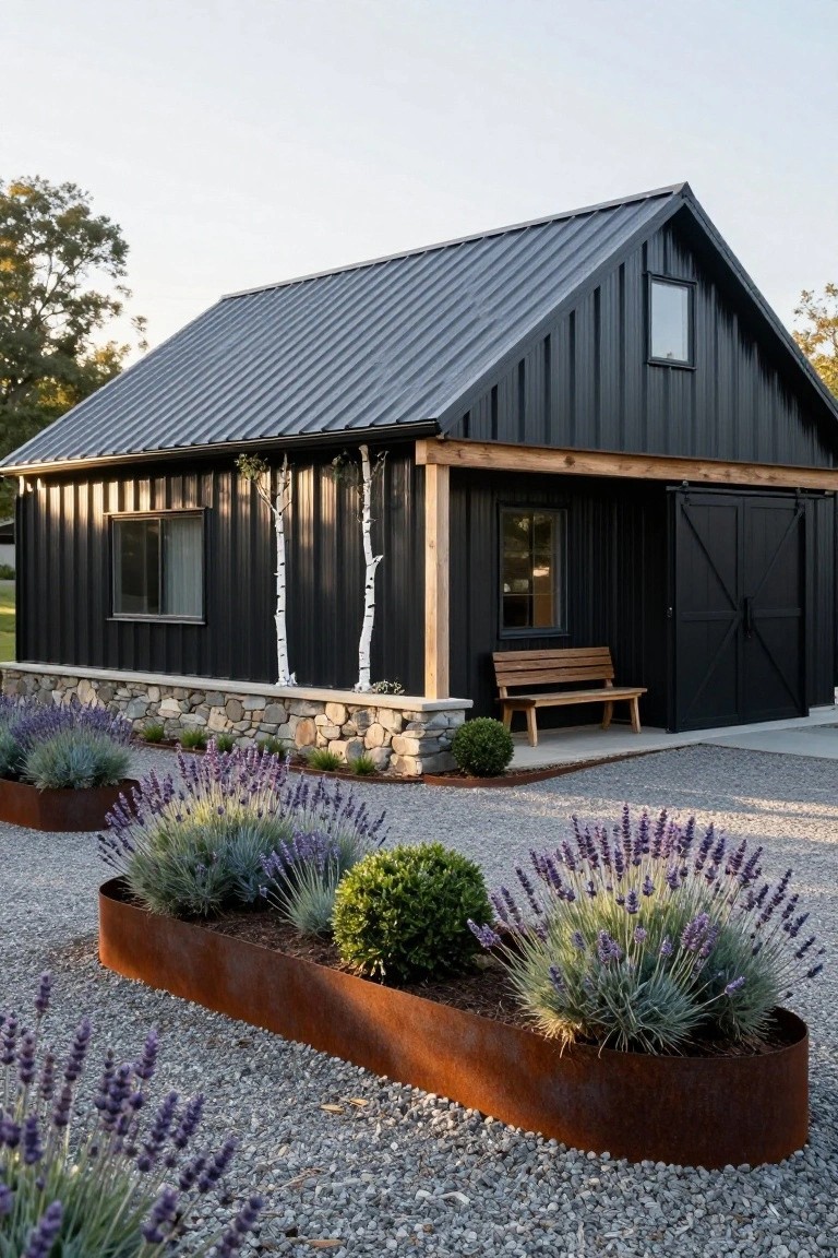 Black metal-sided gabled building with stone foundation, wooden bench, birch trees against the siding, lavender and grasses in curved rusty metal planters, and gravel ground cover.