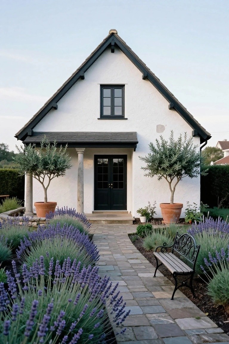 White stucco house with black trim on gabled roof and windows, black double doors under a small porch with columns, flanked by potted olive trees, lavender beds along a stone path, and a black metal bench.
