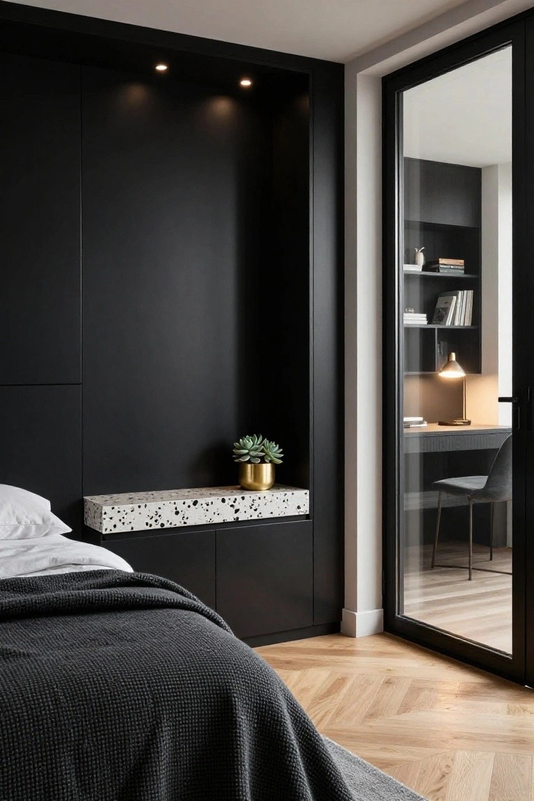 Modern bedroom corner with matte black walls and built-in cabinets next to a bed draped in white sheets and grey blanket, terrazzo shelf with gold plant pot, desk visible through glass door, on herringbone oak floors.