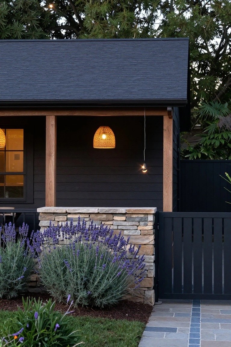 Exterior of a dark-shingled ranch-style house with timber porch posts, rattan pendant light, stone base, lavender bushes, black slatted gate, and surrounding landscaping on a paved path.