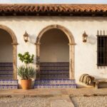 Hacienda-style adobe house exterior with wooden door framed by blue patterned tiles, iron-grilled windows, copper lanterns, potted yucca plant, terracotta steps, and gravel ground.