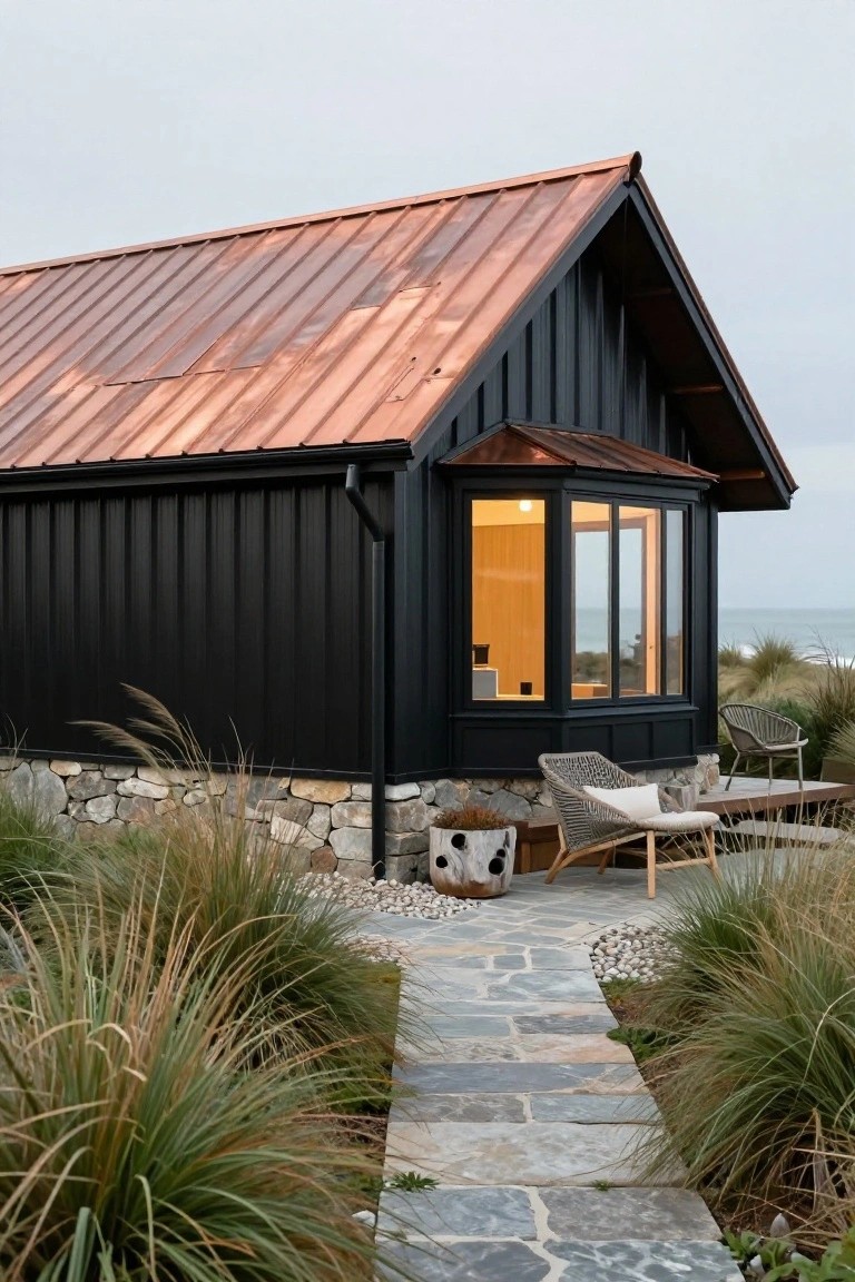 Compact house exterior with black corrugated metal siding, copper roof, stone foundation at base, lit bay window, two woven chairs on wooden deck, stone pathway through grasses near beach and ocean.