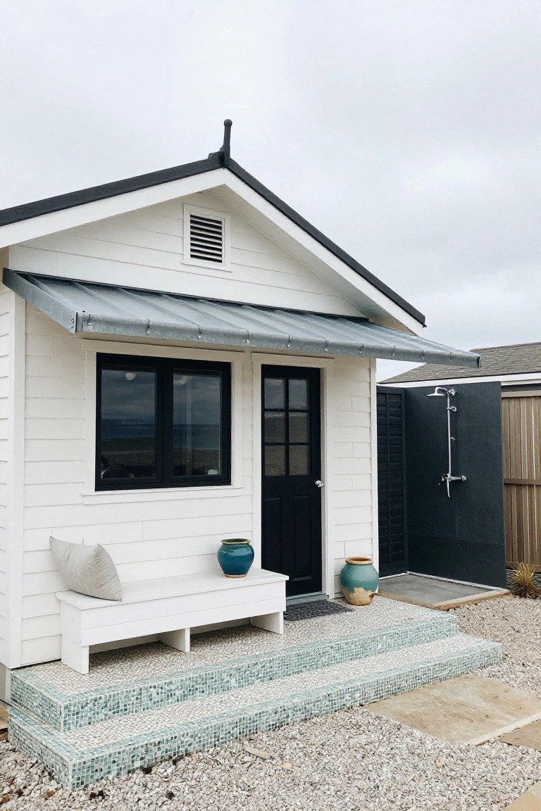 Small white gabled beach cottage with black-framed windows and door, metal awning overhead, white bench with cushion and blue pots beside steps, turquoise mosaic-tiled entry stairs on gravel ground, adjacent black outdoor shower enclosure, and ocean view.