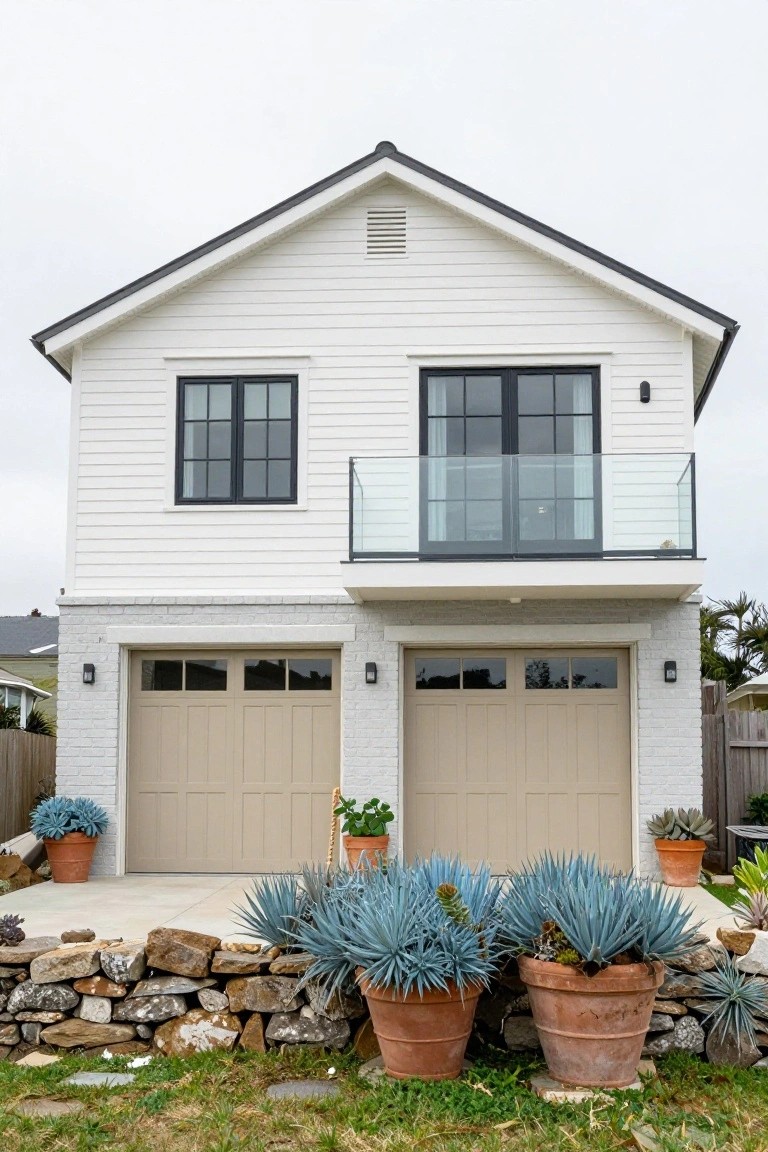 Two-story white shiplap house exterior with black-framed windows, glass balcony above beige double garage doors, stone retaining wall, and large terracotta pots of blue agave plants in the front yard.