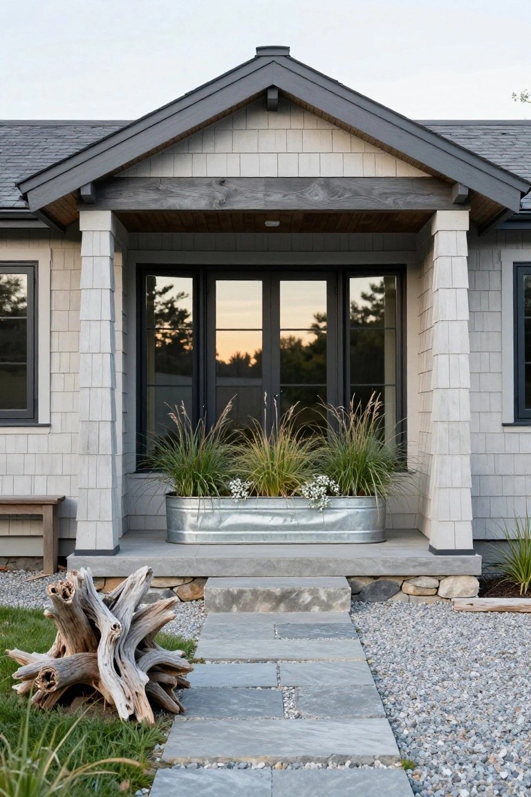 Front exterior of a shingle-style house with gray roof and siding, white columns supporting a gabled covered porch, black-framed glass doors and windows, galvanized metal planter with ornamental grasses and white flowers, concrete steps, flagstone path, gravel ground cover, and driftwood sculpture.