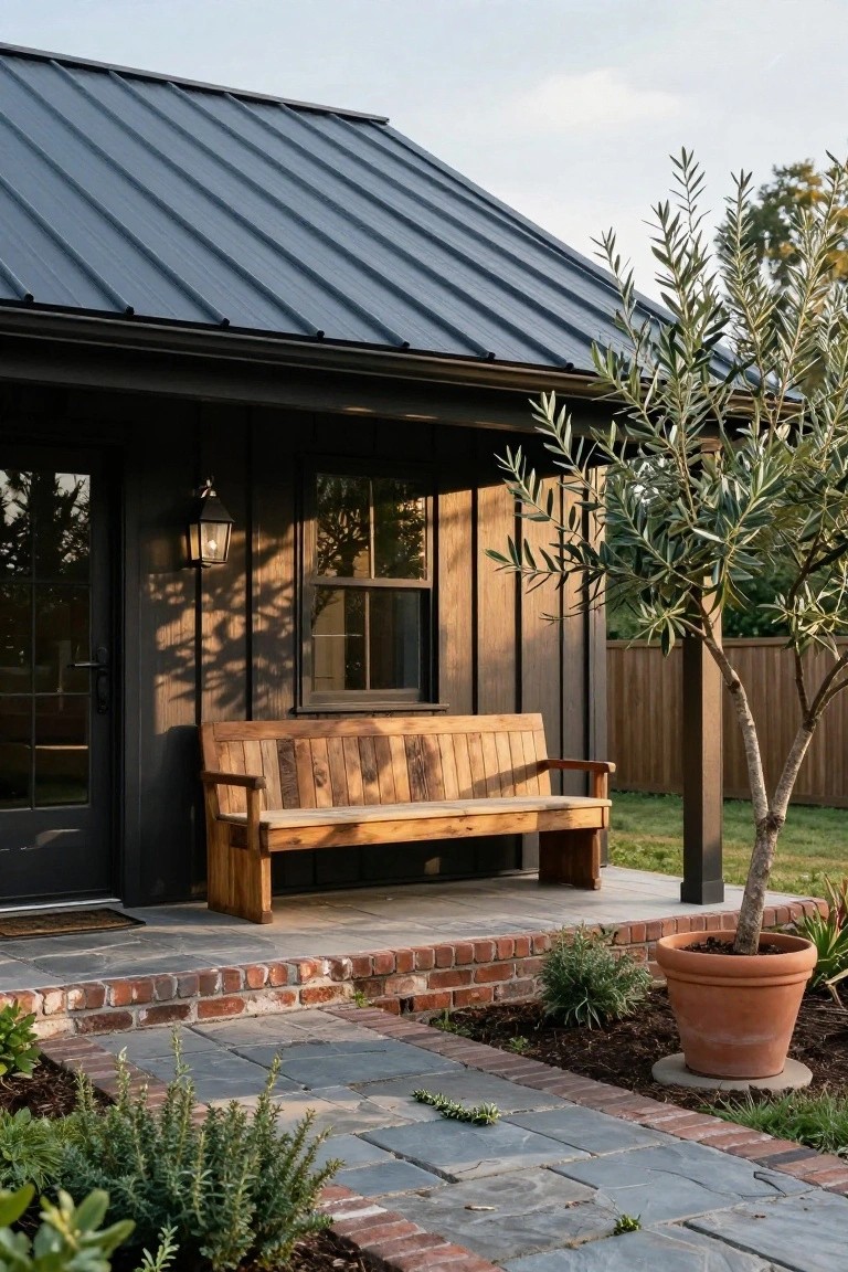 Small dark wood-sided outbuilding with standing-seam dark metal roof, lantern light by glass door and window, wooden bench on brick steps, stone path, potted plants, and olive tree in yard