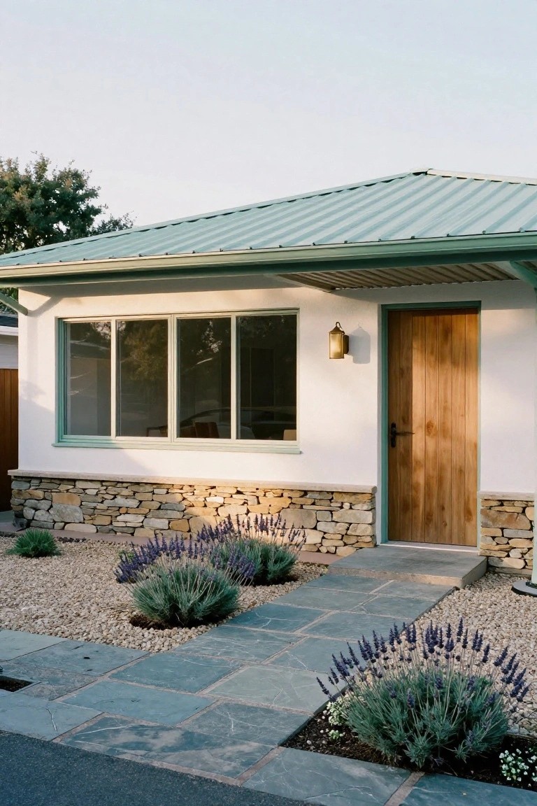 Single-story house exterior with white stucco walls, green standing-seam metal roof, large triple window, wooden plank front door with black handle and brass lantern light, stone base, slate tile entry path, gravel yard with lavender plants.