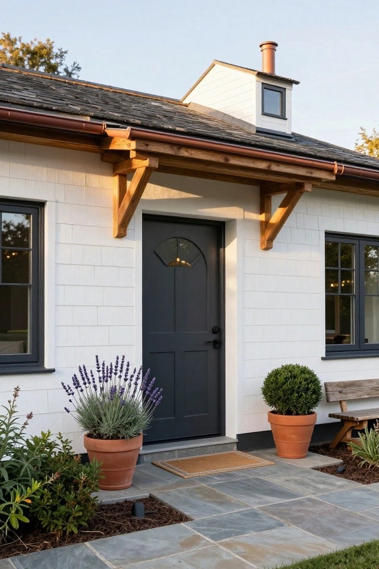 White shiplap-clad cottage exterior featuring a dark arched front door with fanlight, black-framed windows, wooden brackets under the roof overhang, potted lavender and boxwood plants, and a bluestone pathway.