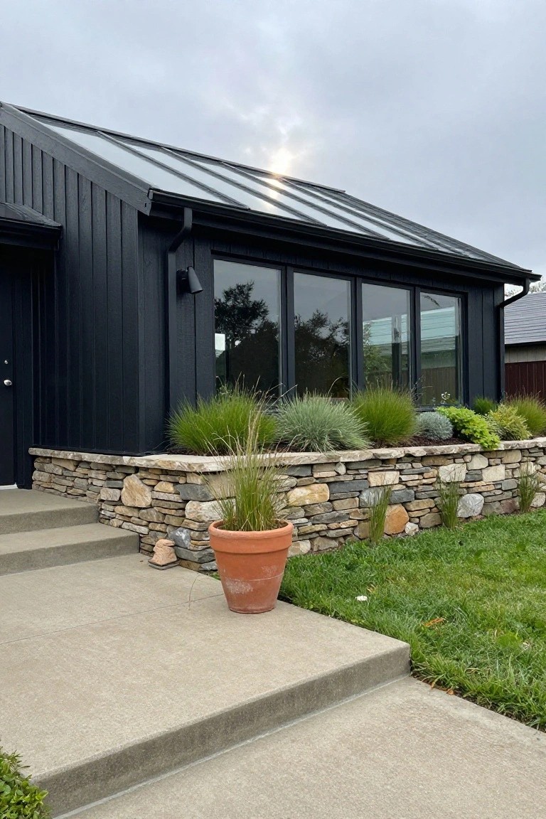 Black board-and-batten sided modern house with sloped glass roof, large glass windows, low irregular stone retaining wall planted with ornamental grasses along the base, concrete entry steps, terracotta planter, and adjacent lawn.