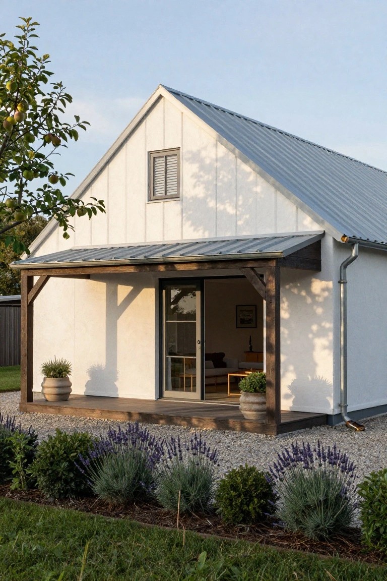 Small gabled white house with standing-seam metal roof, timber-framed covered porch with sliding glass entry door, gravel path edged by lavender plants and potted greenery.