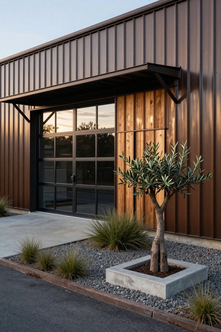 Brown corrugated metal siding on a modern building exterior with a large black-framed glass garage door, vertical wood panels, a potted olive tree in a concrete square planter, grasses, gravel edging, and asphalt driveway.