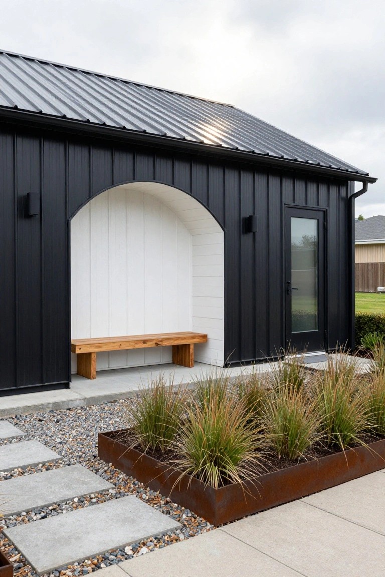 Black metal-sided building with white arched recess containing wooden bench on concrete base, black glass door to the side, wall-mounted lights, gravel path with pavers, and Corten steel planter of ornamental grasses.