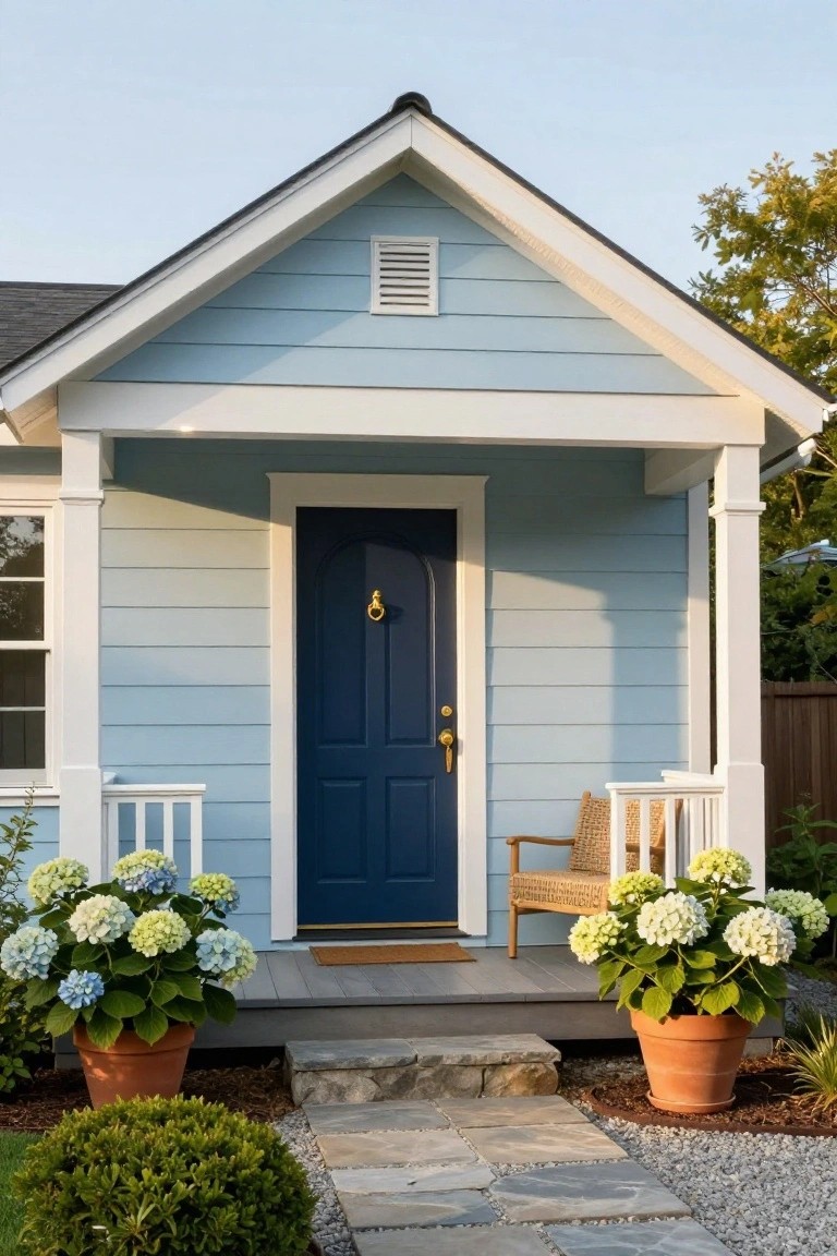 Light blue horizontal clapboard house exterior with white trim, gabled roof, navy arched front door with brass knocker and knob, covered porch with wicker chair, potted hydrangeas flanking steps, stone path, and gravel landscaping.