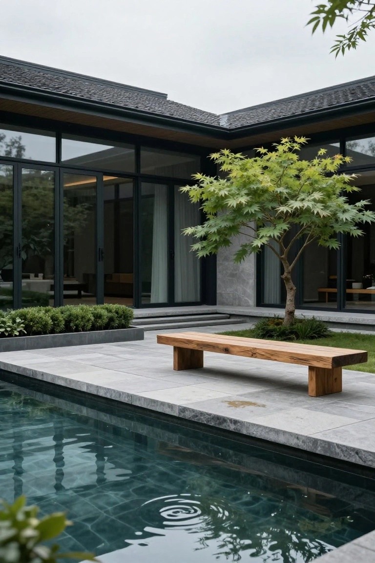 Contemporary house with dark framed glass walls and a wooden bench on gray stone patio beside a rectangular teal pool, featuring a green maple tree and low shrubs.