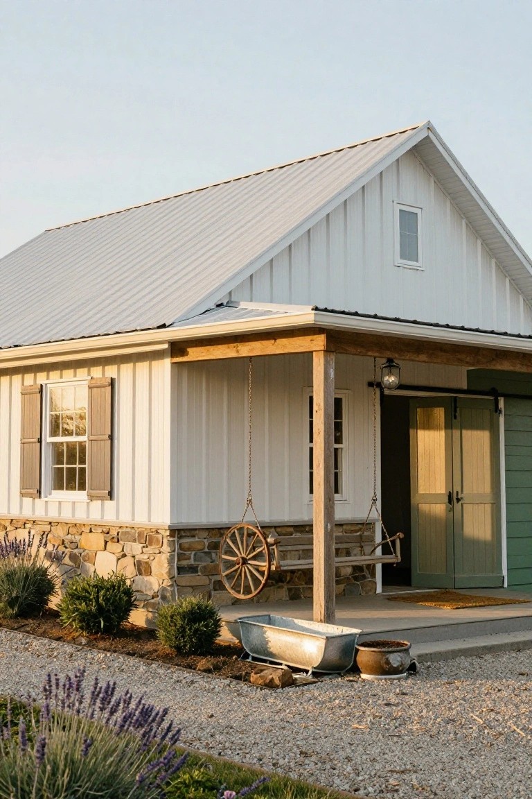 White board-and-batten sided house with metal roof, stone foundation, covered porch featuring a hanging swing and wagon wheel, green double doors, and gravel path with lavender plants.