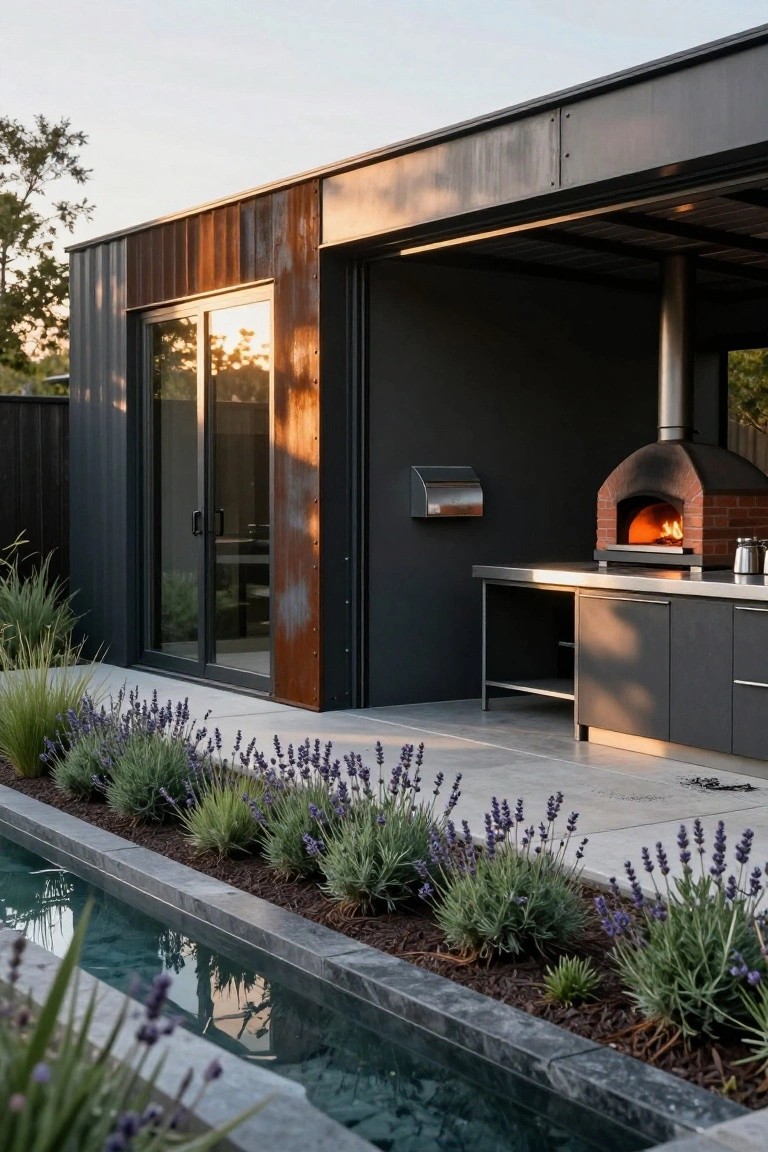 Black metal shipping container-style building with corten steel accents and open sliding glass doors, next to a stainless steel outdoor kitchen counter and brick wood-fired pizza oven with fire inside, lavender plants bordering a narrow stone-edged reflecting pool.