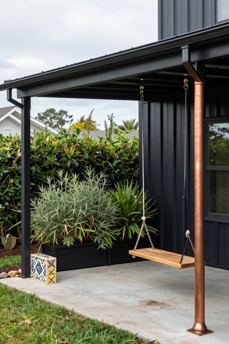 Side view of a black metal-clad house exterior with a covered porch area, wooden bench swing hanging from ropes on a copper pipe post, surrounded by potted plants and hedges on concrete paving.