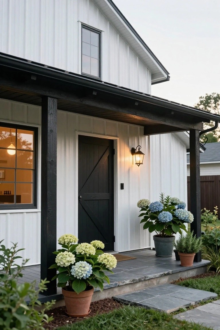 White metal-sided house exterior featuring a black barn door entry under a covered porch with dark wood posts, a wall lantern, potted hydrangeas flanking the steps, and slate stone landing.