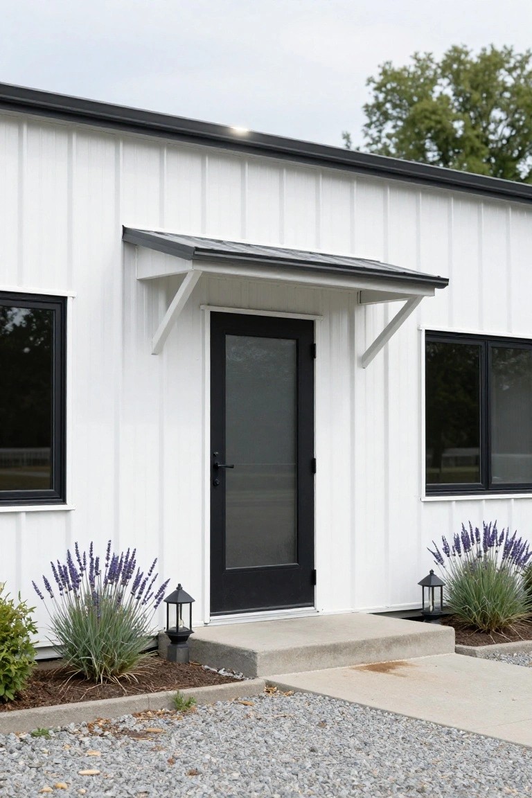 White metal building exterior featuring a black frosted glass front door under a small gray awning, with black-framed windows on either side, lavender plants and lanterns flanking the concrete stoop, and a gravel path in front.