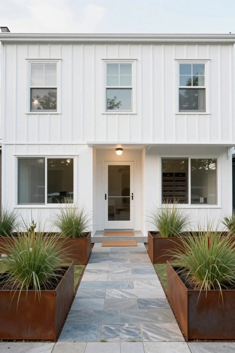 White board-and-batten sided house exterior with vertical siding, three upper windows, side windows, centered white door with glass under small porch light, flanked by rusted metal planters with tall grasses, and gray stone pathway.