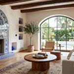Hacienda living room with white stucco walls, exposed wooden beam ceiling, blue-tiled fireplace, beige sofa, rattan chair, wooden coffee table, olive tree, blue vases, and arched black-framed window with outdoor view.