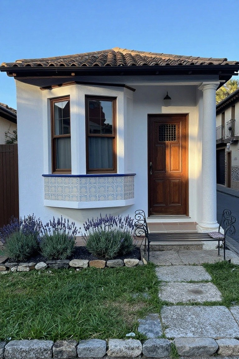 White stucco Spanish-style house exterior with terracotta tile roof, bay window edged in blue tiles, wooden front door with metal grille, single column supporting porch roof with black iron bench, lavender bushes, and stone-edged path.