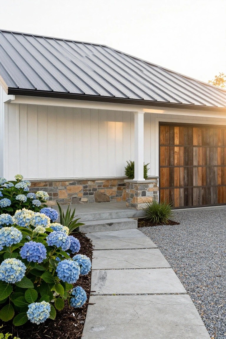 Exterior of a house featuring white board-and-batten siding, dark metal standing-seam roof, stone base at garage with vertical wood-plank garage door, blue hydrangea bushes beside concrete steps and path to gravel driveway.
