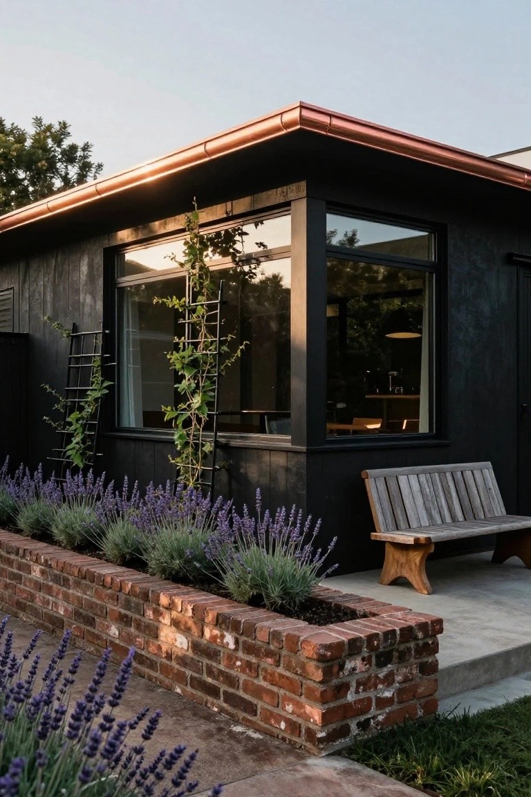 Corner exterior of a black charred wood-clad house with copper roof flashing, large window, vine trellises, lavender bed in brick planter, wooden bench, and concrete steps.