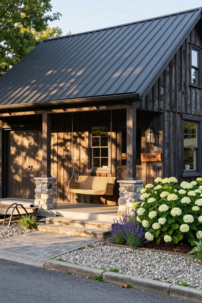 Small gable-roofed cottage with dark vertical wood siding, black standing-seam metal roof, covered porch with woven hanging swing between stone pillars, hydrangea bushes, gravel beds, and adjacent road.