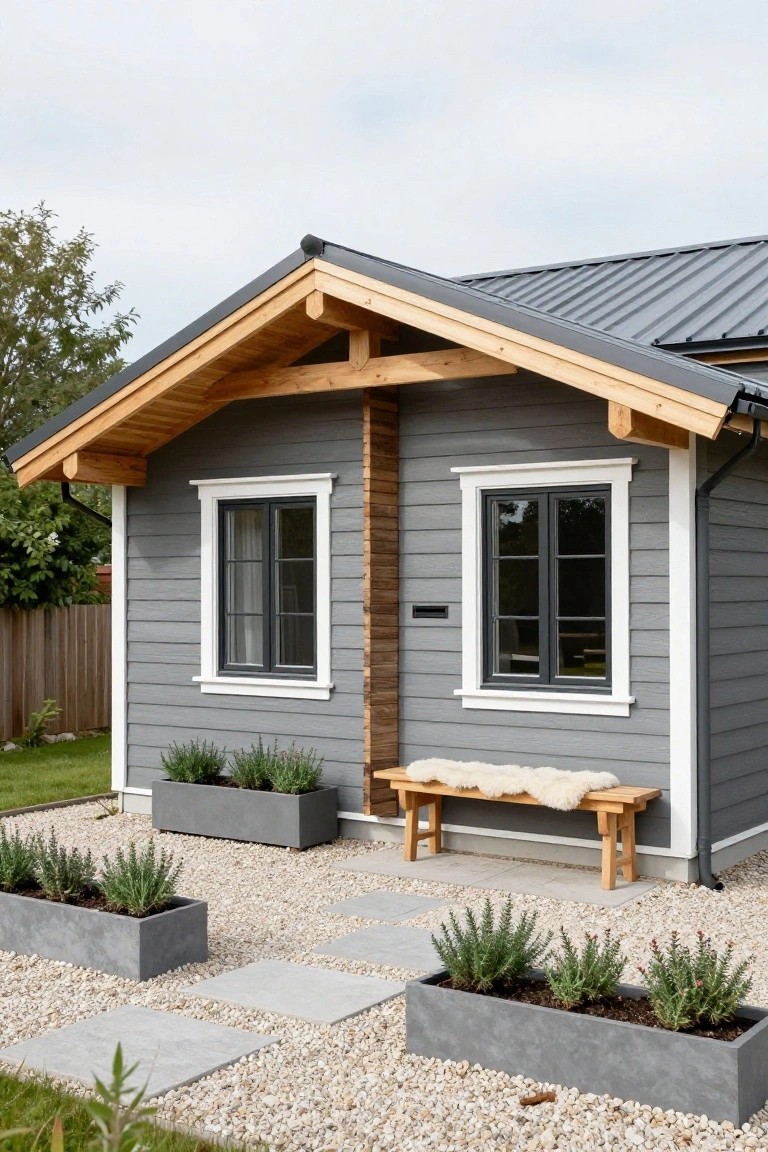 Small gray-sided cabin with exposed wooden beams and posts, black-framed windows, dark metal roof, wooden bench with sheepskin, potted herbs, gravel ground, and stone pavers in front.