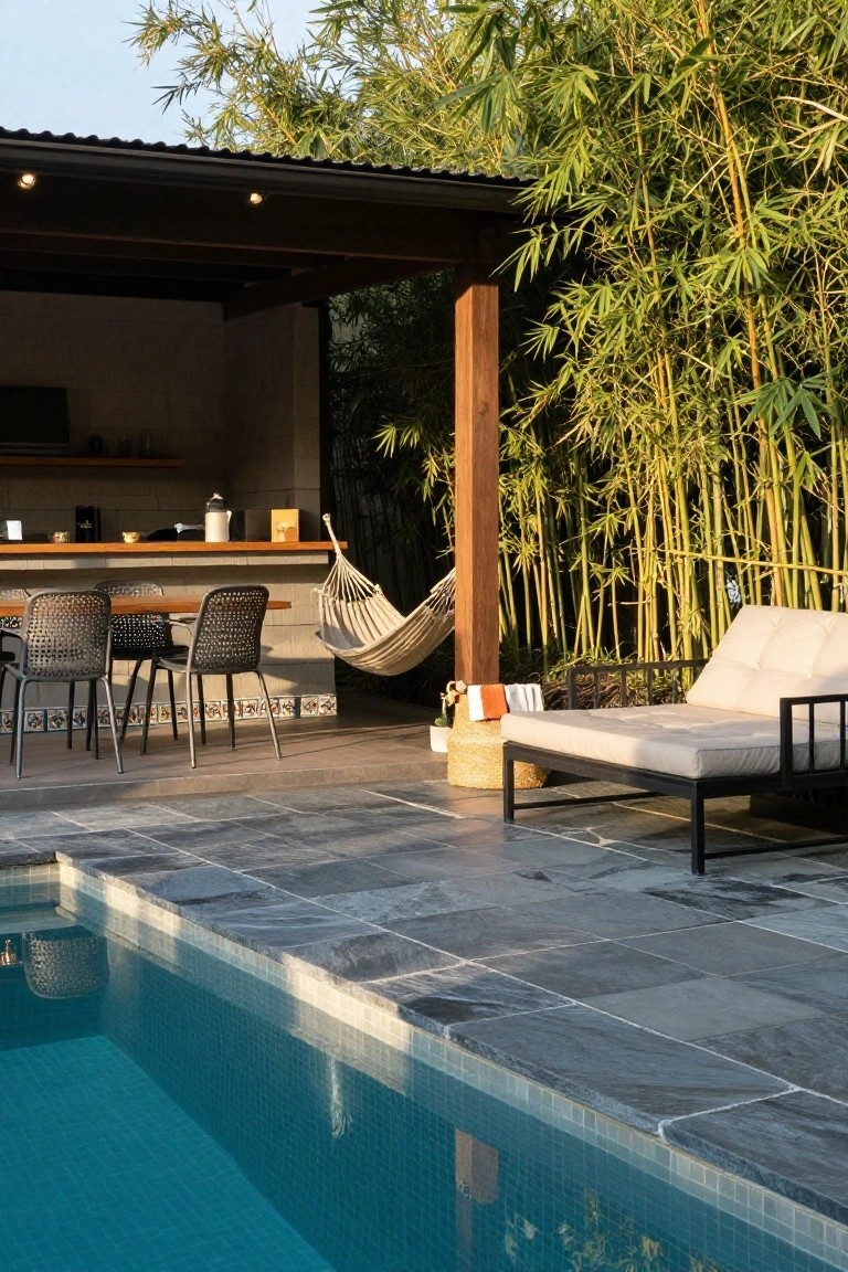 Outdoor pool area with a covered bar counter, stools, hammock, daybed, and tall bamboo screening beside a tiled pool deck.