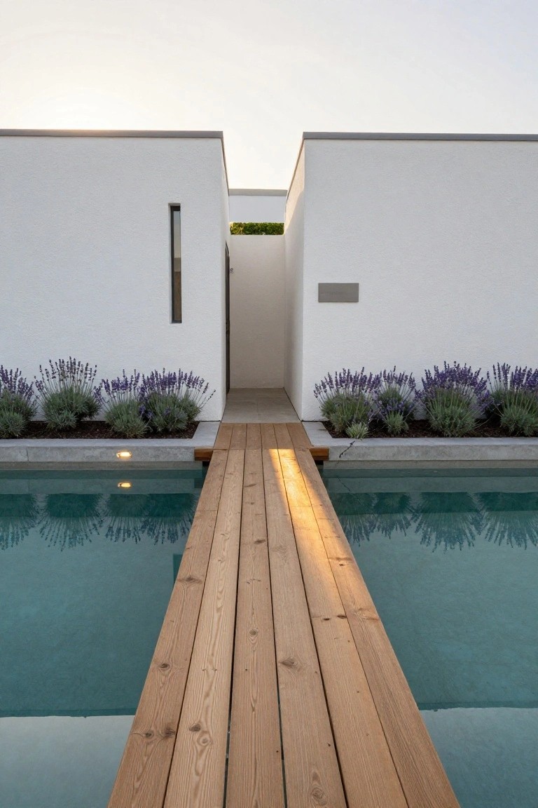White minimalist flat-roofed house exterior with a wooden bridge walkway spanning a narrow rectangular pool, bordered by lavender shrubs and gravel beds, leading to a recessed entry door.