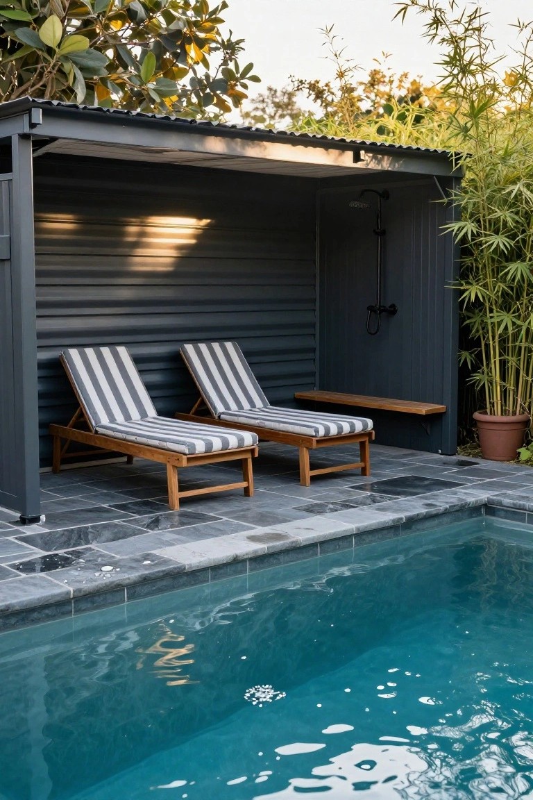 Gray metal-roofed outdoor shelter with two beige-and-white striped wooden lounge chairs, wooden bench, and black shower head beside a turquoise swimming pool on dark slate tiles, surrounded by bamboo plants and greenery.