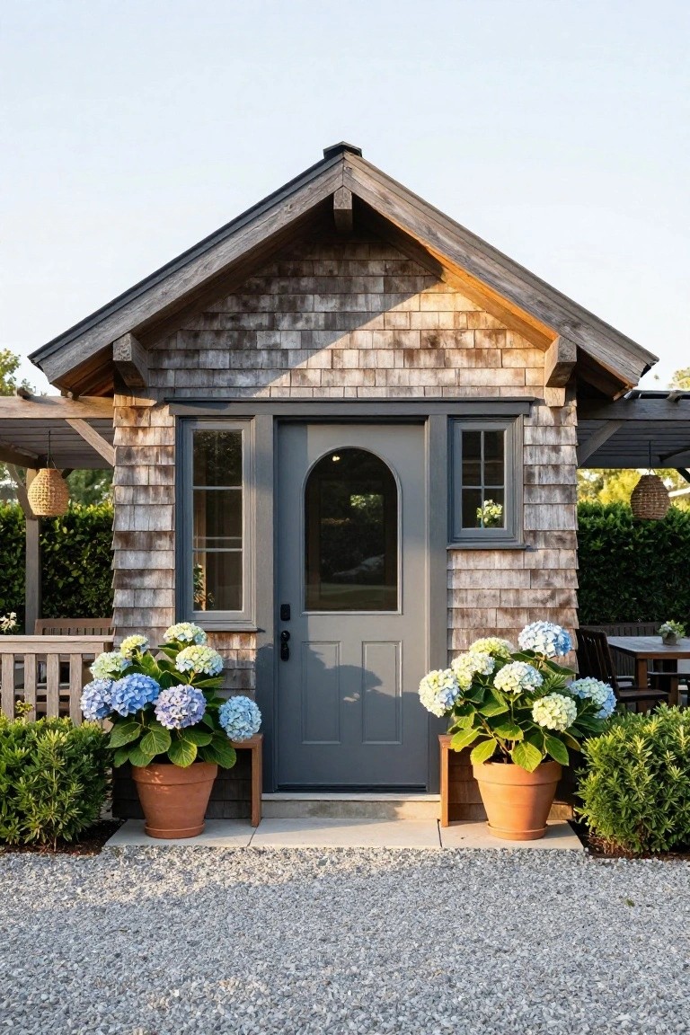Small shingled cottage with gray front door and arched window, flanked by two large terracotta pots filled with blue hydrangea blooms, gravel ground, and boxwood shrubs nearby.