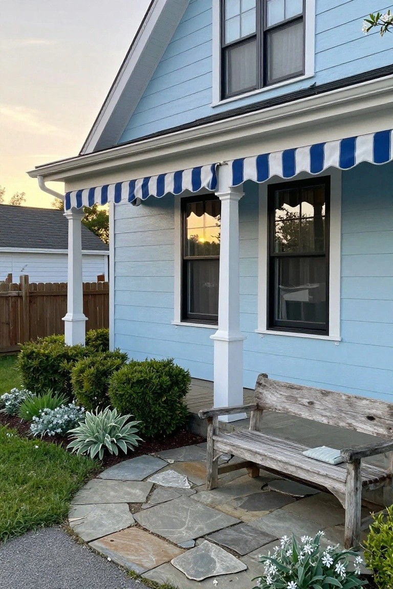 Side exterior of a light blue clapboard house featuring a covered porch with blue-and-white striped awning, white columns, dark-framed windows, boxwood shrubs, stone pathway, and weathered wooden bench with pillow and book.