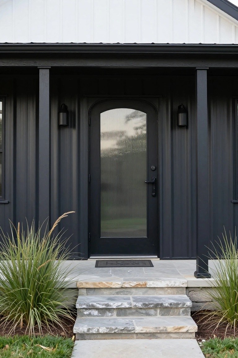 Front view of a small house with black corrugated metal siding, black-framed arched glass entry door on a covered porch supported by columns, stone steps, wall lanterns, and tall ornamental grasses on either side.