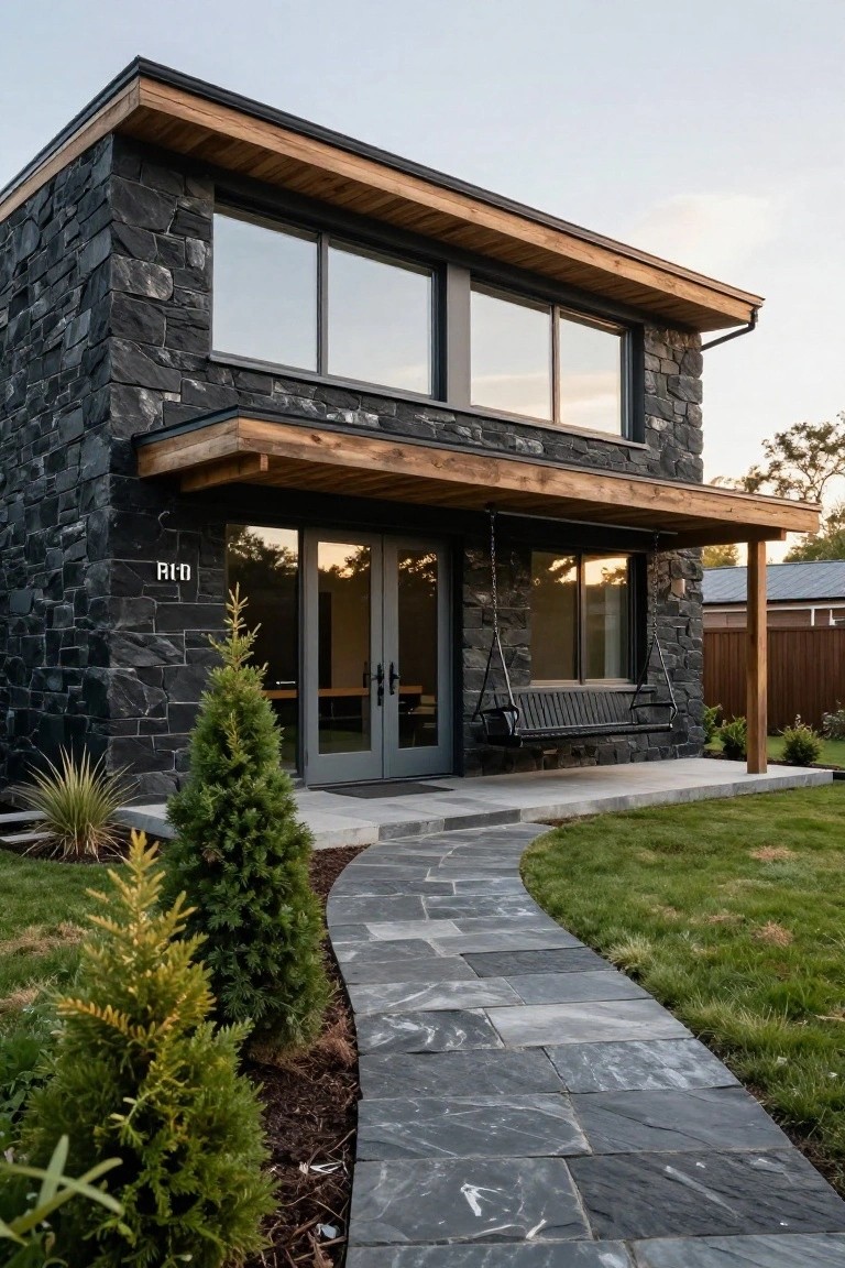 Modern cottage exterior with black stone walls, wooden roof overhang and porch, large glass windows and doors, hanging porch swing, curved stone pathway, and small evergreen shrubs in the front yard.