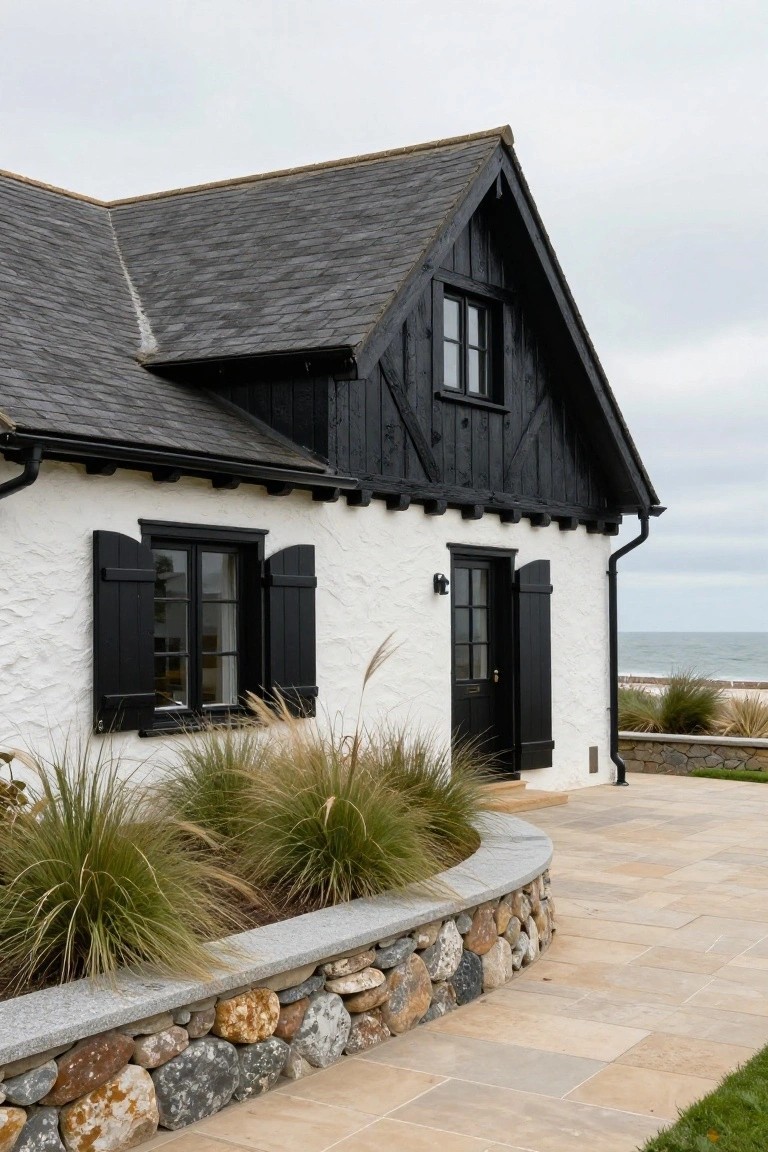White stucco house with black timber framing, black-framed windows and shutters, black front door, dark slate roof, low stone wall with grasses, and paved path near beach dunes.
