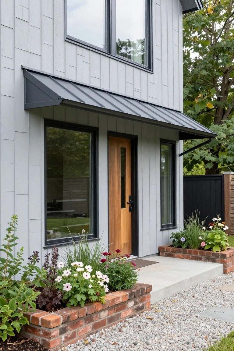Gray board-and-batten house exterior with black-framed windows, oak entry door under angled black metal awning, brick planters filled with flowers and greenery flanking a concrete stoop and gravel path.