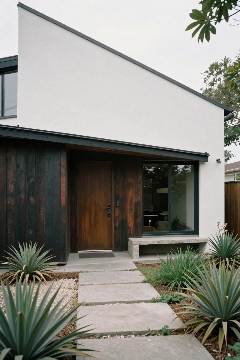 Modern house facade with white stucco walls, angled black roofline, dark vertical wood cladding beside a wooden entry door, concrete bench, stone pathway, and agave plants in gravel beds.
