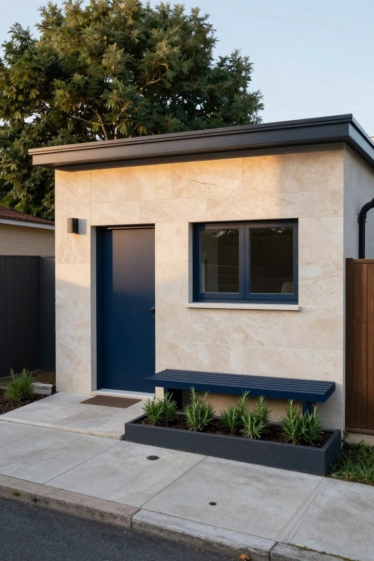 Small modern home exterior with pale beige stone tile cladding, navy blue front door, single dark-framed window, wall light, blue bench, black planter box with plants, concrete path, and surrounding fences and trees.