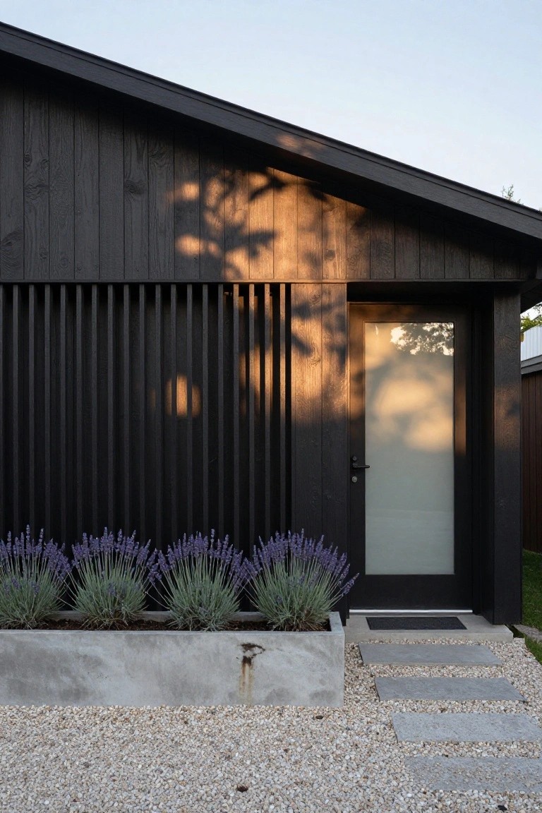 Modern house exterior with black vertical wood slats beside a frosted glass entry door, lavender plants in a concrete planter box, gravel ground cover, and stone steps.