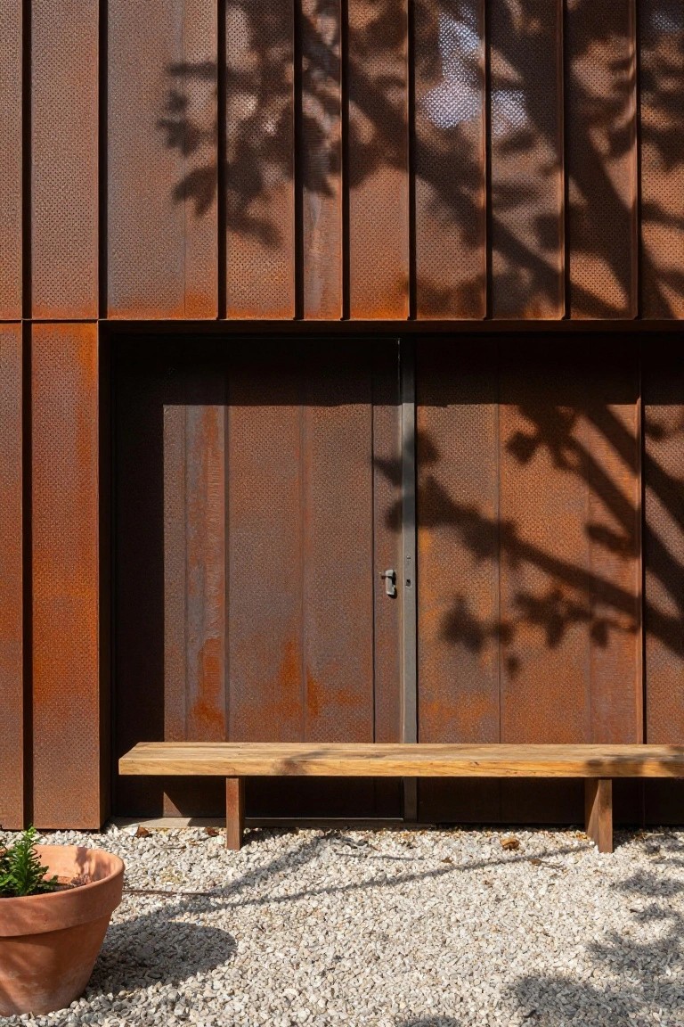 Vertical perforated rusted metal siding on a house exterior with a dark metal double door, wooden bench, gravel ground, and terracotta pot with small plant nearby.