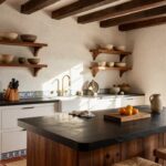 Hacienda-style kitchen interior with black marble-topped island, white cabinets, rattan-wrapped wooden bar stools, terracotta tile floor, arched black-framed window overlooking garden, wooden open shelves with pottery, and blue tiled backsplash.